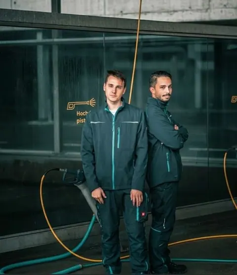Zwei Männer in dunklen Arbeitsjacken vor einer modernen Werkstatt mit professionellem Equipment